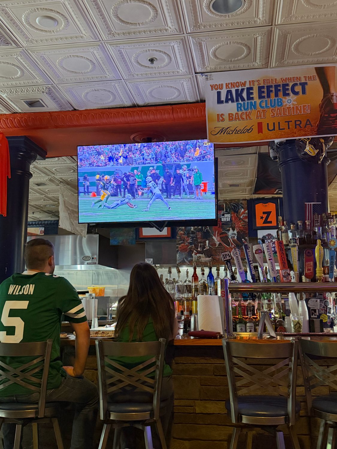 Two fans in Jets jerseys at the bar watching the game on the big screen